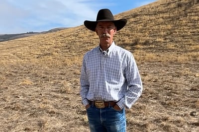 A man wearing a cowboy hat stands with his hands in his pockets on a dry, grassy open space area.