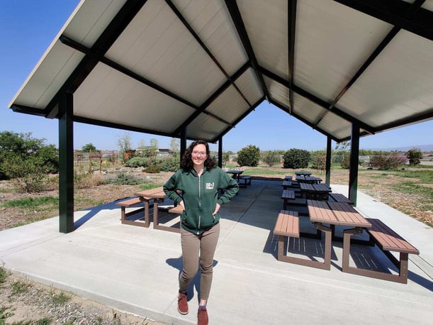 Our City Forest employee in green sweatshirt smiling and standing in front of outdoor pavilion with picnic tables
