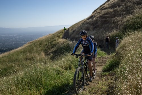 Mountain bikers riding towards the camera on a dirt path wrapping around a hillside with green and gold grass.